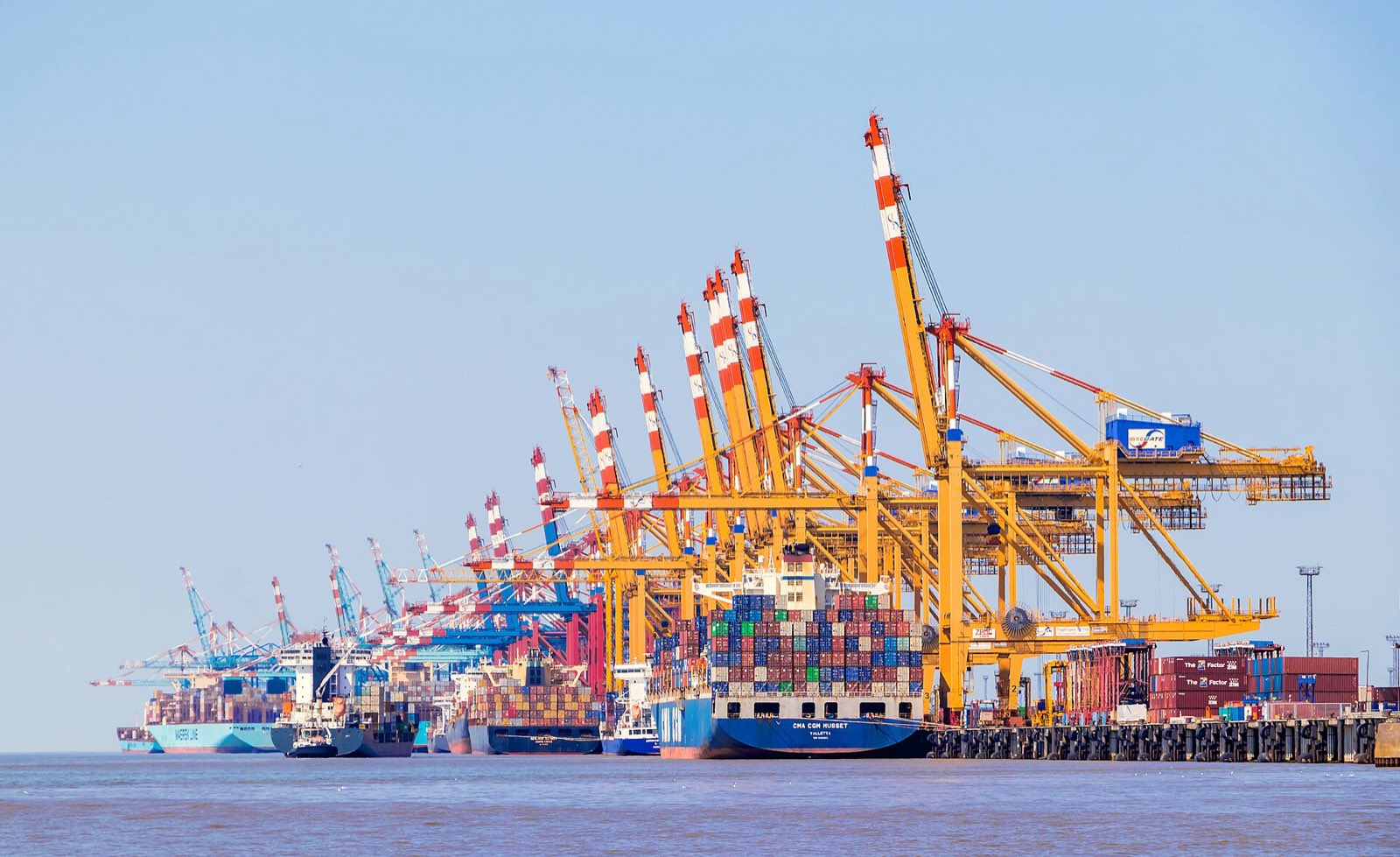 Stacked shipping containers at a container port, with gantry cranes behind.